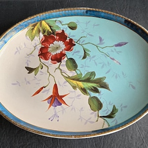May include: Oval serving tray with a floral design. The tray features a light blue and cream background with a vibrant floral pattern in red, green, and purple. The rim is trimmed with gold and blue accents, and it has handles on each side.