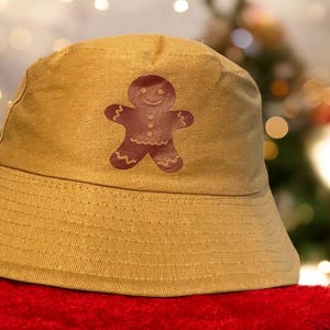 May include: A tan bucket hat with a brown gingerbread man design. The hat has a wide brim and a flat crown.