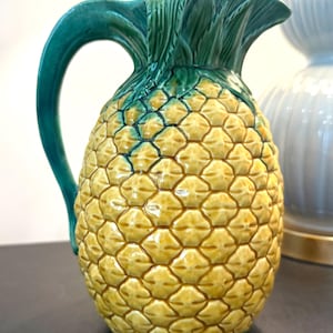 May include: A ceramic pitcher shaped like a pineapple with a yellow textured body and a green handle and top leaves. The surface features a repeating diamond pattern across the yellow section, while the green handle curves upward to form the spout.