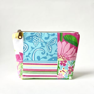 May include: A rectangular fabric pouch with a gold-tone metal zipper closure. The exterior features a patchwork design with light blue panels showing white fish, pink floral patterns, and horizontal stripes in pink, yellow, and green.