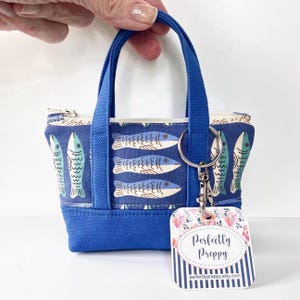 May include: A small blue tote bag with a fish pattern and blue handles. The bag has a zipper closure and a keychain attached. A tag with the text "Perfectly Preppy" hangs from the keychain.