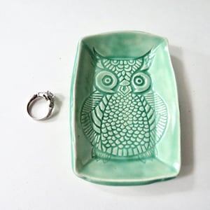 May include: A light green ceramic owl-shaped ring dish with a detailed owl design. A silver ring with a square stone sits next to the dish. The dish is rectangular with rounded corners.