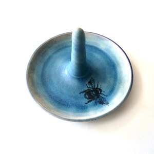 May include: A circular ceramic ring holder with a central cone, featuring a blue gradient glaze and a black bee illustration printed on the flat surface. The dish has a smooth finish and a slightly raised rim, providing a stable base for jewelry.