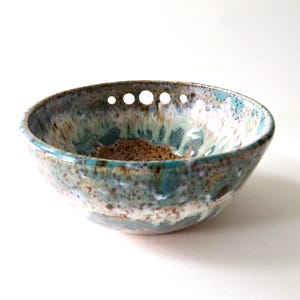 May include: A handmade ceramic bowl with a speckled, glazed finish. The bowl features a blend of blue, green, and brown hues, with a series of small, evenly spaced holes near the rim. The interior has a textured, earthy appearance.