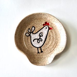 May include: A speckled ceramic spoon rest with a whimsical chicken design. The chicken is white with black outlines and a red comb. The spoon rest is beige with brown speckles and a curved shape.