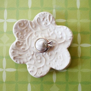 May include: A white, flower-shaped ceramic ring dish with an ornate, embossed design. A silver ring with a clear gemstone rests in the center. The dish sits on a green background with a white floral pattern.