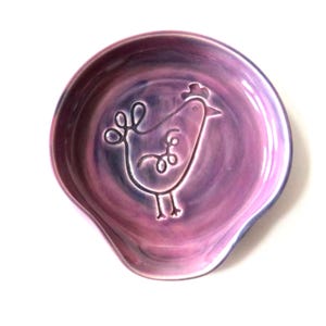 May include: A ceramic spoon rest in shades of purple and lavender, featuring a raised outline of a whimsical chicken. The spoon rest has a curved shape with a small lip for resting utensils. The background is white.