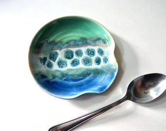 Puede incluir: Un reposacucharas de cerámica con un esmalte azul, verde azulado y blanco. El reposacucharas tiene una forma única con un borde curvo y un patrón texturizado. Una cuchara plateada descansa junto al reposacucharas.