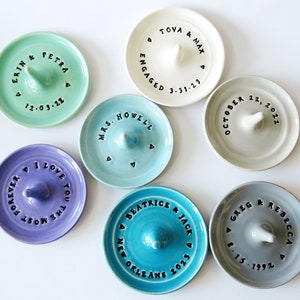 May include: Six ceramic ring dishes with various colors and personalized messages. The messages include names, dates, and phrases like "engaged" and "I love you the most forever".