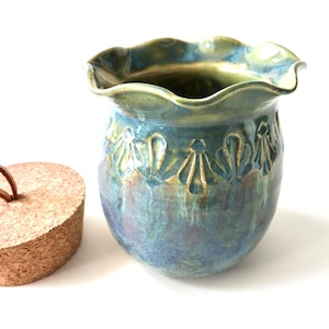 May include: A handcrafted ceramic vase with a ruffled rim, glazed in shades of blue and green. The vase features a floral pattern and is accompanied by a cork stopper with a leather loop. A decorative piece for the home.