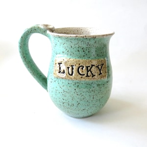 May include: A speckled turquoise ceramic mug with a curved handle. The mug features the word "LUCKY" in raised letters on a rectangular beige patch. The interior rim is a creamy white color.