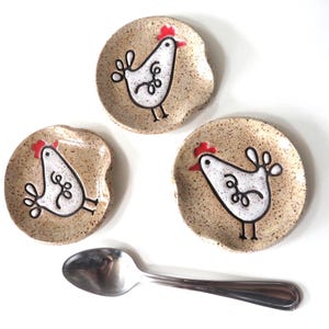 May include: Three speckled ceramic spoon rests with a whimsical chicken design in black, white, and red. A stainless steel spoon rests below the spoon rests. The spoon rests are beige with a speckled pattern.