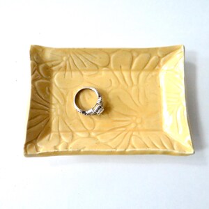 May include: A rectangular, light yellow ceramic ring dish with a floral pattern. A silver ring with a large, clear gemstone sits in the center. The dish has slightly curved edges.