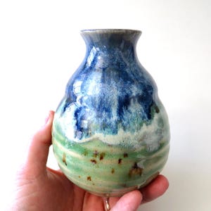 May include: A handmade ceramic vase with a rounded body and a narrow neck. The vase features a gradient of colors, transitioning from green at the base to blue at the top. The vase is held by a person.