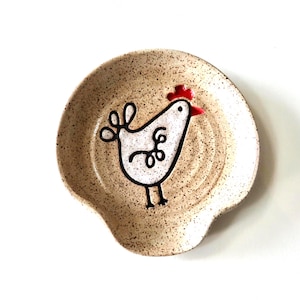 May include: A speckled ceramic spoon rest with a whimsical chicken design. The chicken is white with black outlines and a red comb. The spoon rest is beige with a curved shape and a small indentation.
