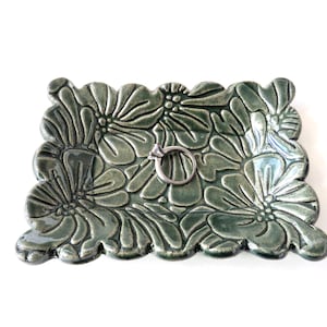 May include: A rectangular, ceramic ring dish in a muted green hue, adorned with a floral pattern. The dish features a raised, textured design of stylized flowers and holds a silver ring. The edges of the dish are scalloped.