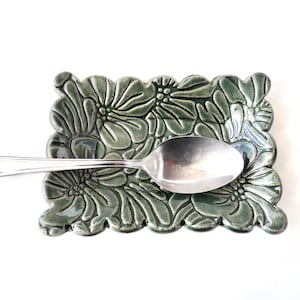 May include: A ceramic spoon rest in a rectangular shape with a floral design in shades of green. A silver spoon rests on the spoon rest. The spoon rest is a decorative kitchen accessory.