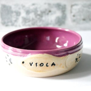 May include: A ceramic pet bowl with a purple interior and rim, and a cream-colored exterior. The name "Viola" is written in black letters on the bowl, with paw prints on either side. The bowl is designed for pets.