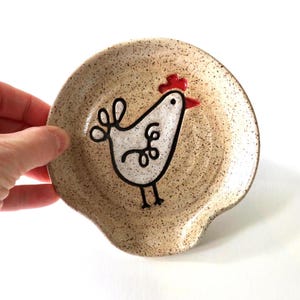 May include: A round ceramic spoon rest with a speckled beige finish. The center features a hand-painted white chicken illustration with a red comb and beak, outlined in black, with a curved handle rest at the bottom edge of the dish.