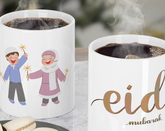 Op de afbeelding: Twee witte keramische mokken met feestelijke ontwerpen. Een mok heeft een cartoonillustratie van twee kinderen die vuurwerk vasthouden, terwijl de andere mok de tekst "eid mubarak" in gouden schrift heeft.