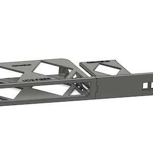 Puede incluir: Una estructura geométrica gris con espacios abiertos y el texto "UCO-FIBER". El diseño incluye recortes rectangulares y triangulares, y la forma general es alargada con una serie de aberturas cuadradas en un lado. La estructura parece estar impresa en 3D.