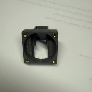 Pode incluir: Um componente de pl&aacute;stico cinza escuro, impresso em 3D, com uma parte superior quadrada e uma abertura circular. Quatro pequenos fixadores dourados s&atilde;o vis&iacute;veis ao redor da abertura circular. O componente parece ser um suporte.