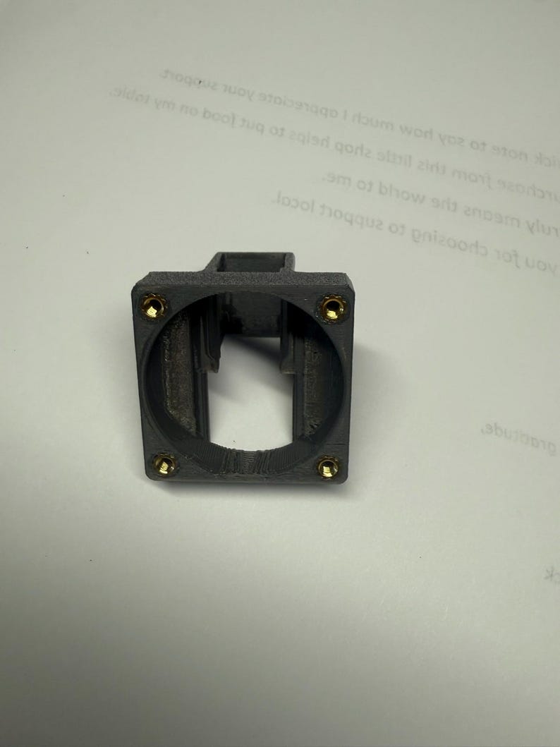 Pode incluir: Um componente de pl&aacute;stico cinza escuro, impresso em 3D, com uma parte superior quadrada e uma abertura circular. Quatro pequenos fixadores dourados s&atilde;o vis&iacute;veis ao redor da abertura circular. O componente parece ser um suporte.