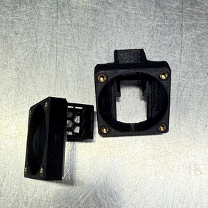 Pode incluir: Componentes pretos impressos em 3D com parafusos cor de lat&atilde;o. As pe&ccedil;as incluem uma caixa quadrada com uma abertura retangular, uma caixa de ventilador menor e uma grelha em favo de mel. Os itens est&atilde;o dispostos em uma superf&iacute;cie met&aacute;lica.
