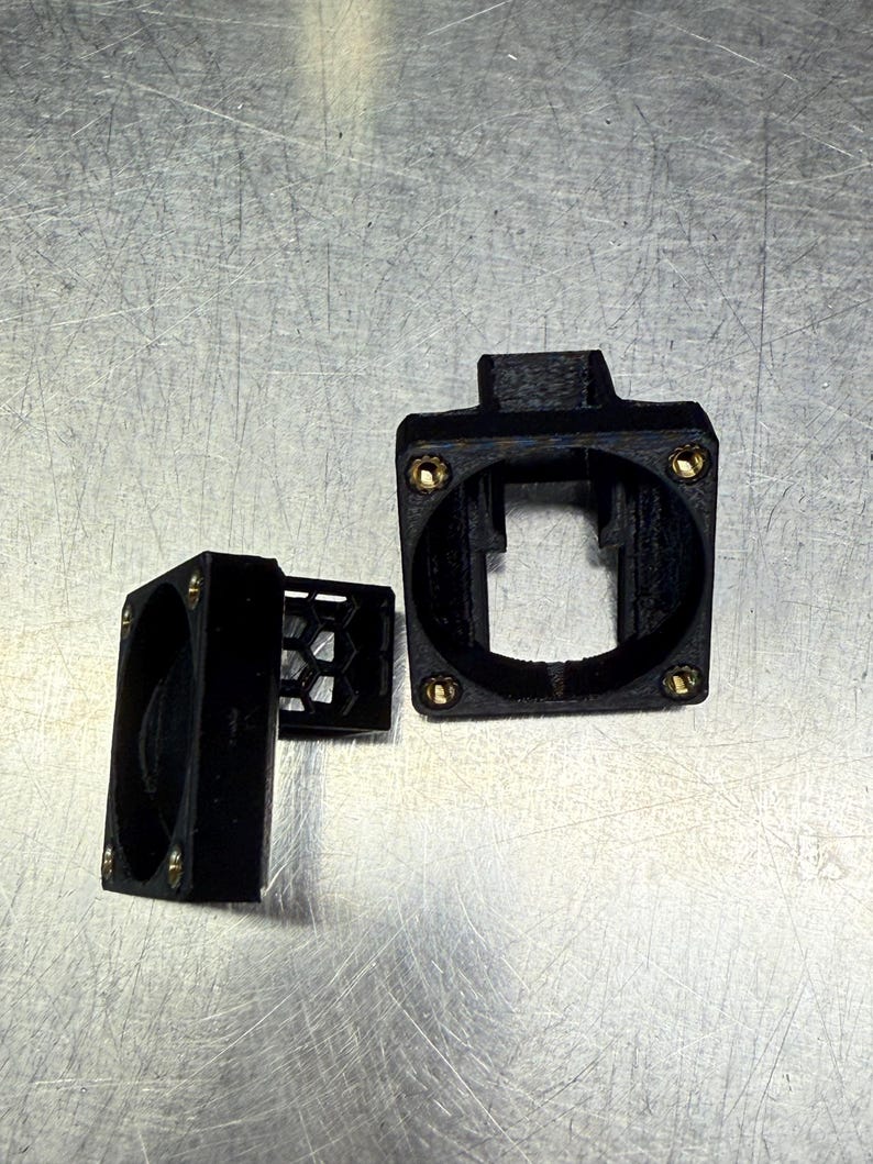 Pode incluir: Componentes pretos impressos em 3D com parafusos cor de lat&atilde;o. As pe&ccedil;as incluem uma caixa quadrada com uma abertura retangular, uma caixa de ventilador menor e uma grelha em favo de mel. Os itens est&atilde;o dispostos em uma superf&iacute;cie met&aacute;lica.