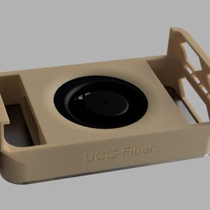 May include: A beige rectangular device featuring a central black circular element. The device has raised sides and the text "UCC-Fiber" is visible on the front. The design includes cut-out shapes on the sides.