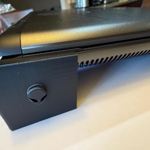 May include: Black laptop computer resting on a black stand. The stand has a circular emblem with an alien head design. The laptop has a vent with multiple angled slots.