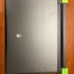 May include: A dark gray laptop computer with a black screen and a green alien logo on the lid. The laptop is propped up on two green plastic stands.