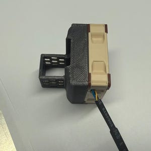 Pode incluir: Um conector el&eacute;trico impresso em 3D com uma caixa preta e bege. O conector tem uma forma retangular com ranhuras e um fio com isolamento azul, amarelo e preto. O fio est&aacute; conectado &agrave; parte bege do conector.