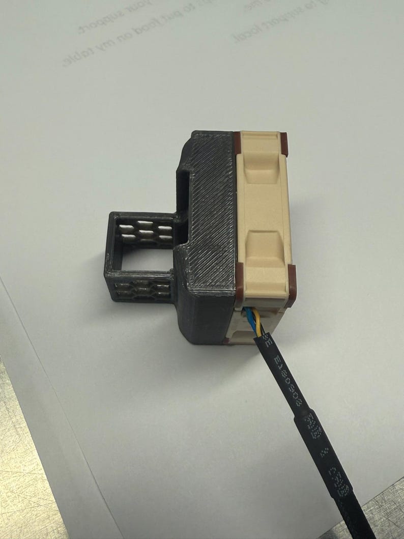 Pode incluir: Um conector el&eacute;trico impresso em 3D com uma caixa preta e bege. O conector tem uma forma retangular com ranhuras e um fio com isolamento azul, amarelo e preto. O fio est&aacute; conectado &agrave; parte bege do conector.