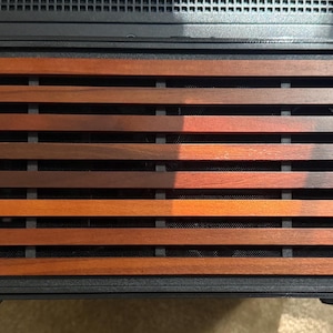 May include: A close-up of a black electronic device with a wooden slat design. The wooden slats are various shades of brown and orange, creating a warm, natural aesthetic. The device has a rectangular shape.