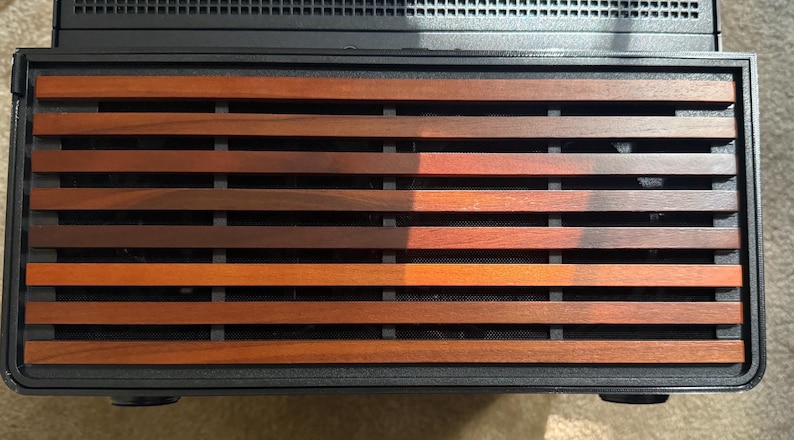 May include: A close-up of a black electronic device with a wooden slat design. The wooden slats are various shades of brown and orange, creating a warm, natural aesthetic. The device has a rectangular shape.