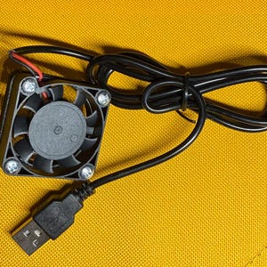 May include: A small, black USB-powered fan with a square frame and a circular fan blade. The fan has a black cable and a USB connector. The fan is on a yellow surface.