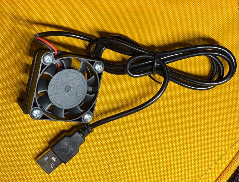 May include: A small, black USB-powered fan with a square frame and a circular fan blade. The fan has a black cable and a USB connector. The fan is on a yellow surface.