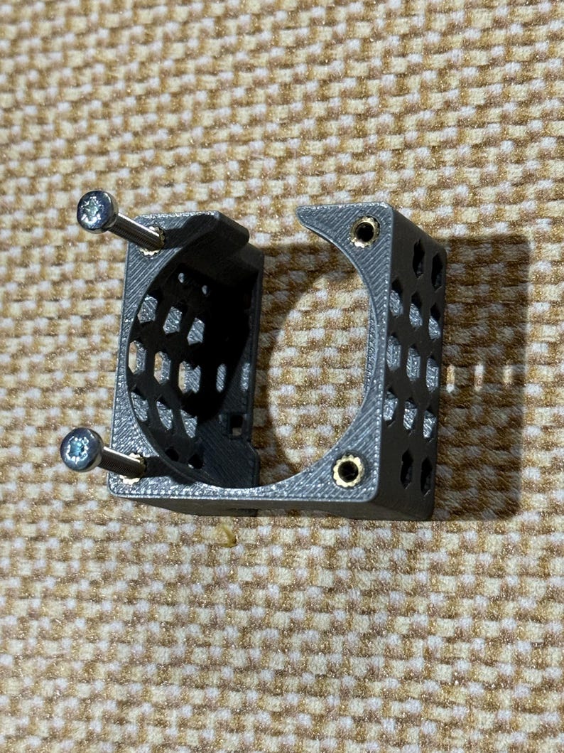 May include: A gray 3D-printed bracket with a semi-circular design and multiple small holes. Two silver screws are inserted through the bracket. The bracket is designed to hold an object, with a curved section and a rectangular section.