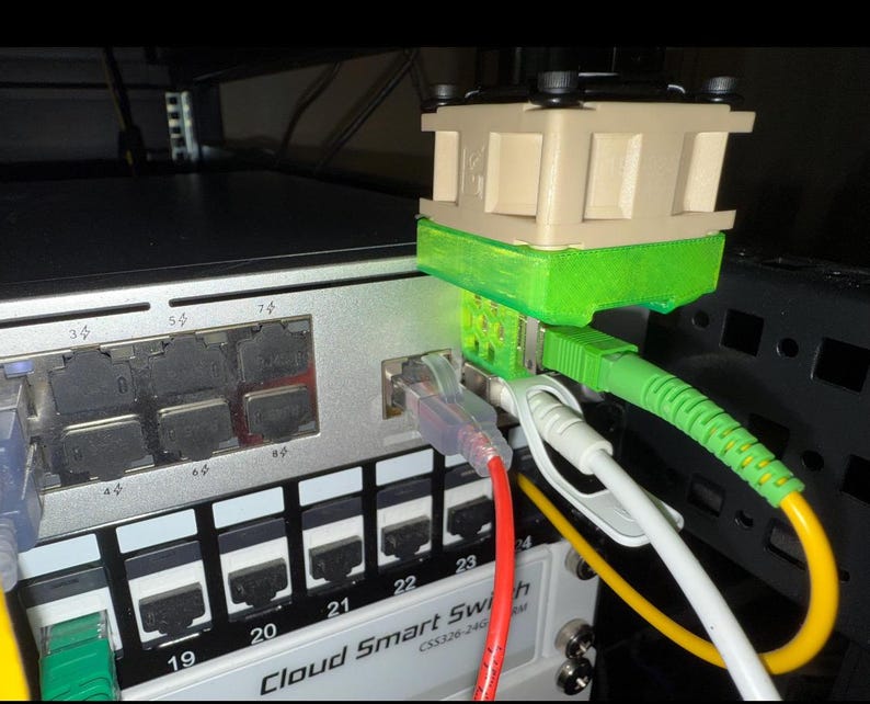 Pode incluir: Close-up de um switch de rede com v&aacute;rios cabos coloridos conectados. Um dispositivo bege e verde est&aacute; conectado ao switch. Os cabos incluem verde, amarelo, branco e vermelho, criando uma configura&ccedil;&atilde;o de rede colorida e organizada.