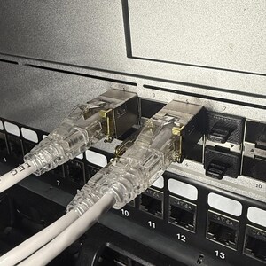 以下が含まれることがあります： 6番と7番のポートに白いイーサネットケーブルが2本接続されたネットワークスイッチのクローズアップ。ケーブルはRJ-45コネクタに接続されています。スイッチは、番号付きのポートを持つ黒い前面プレートを持っています。