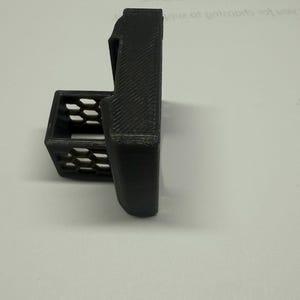 Pode incluir: Um suporte preto impresso em 3D com um corpo retangular e uma pequena cesta aberta. O suporte tem uma superf&iacute;cie texturizada e &eacute; projetado para segurar um item. A cesta tem um padr&atilde;o de favo de mel.