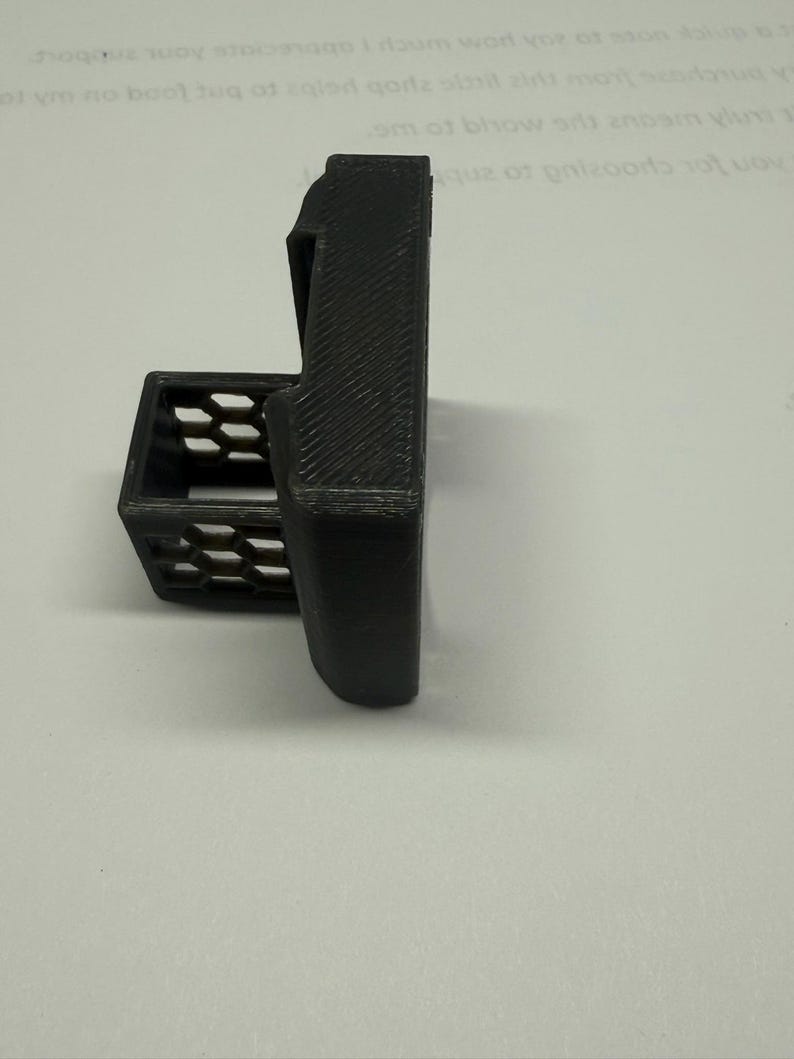 Pode incluir: Um suporte preto impresso em 3D com um corpo retangular e uma pequena cesta aberta. O suporte tem uma superf&iacute;cie texturizada e &eacute; projetado para segurar um item. A cesta tem um padr&atilde;o de favo de mel.