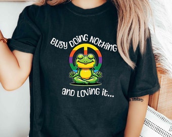 May include: Black t-shirt with a green frog meditating inside a rainbow peace sign. The text "Busy doing nothing and loving it..." is printed on the shirt.