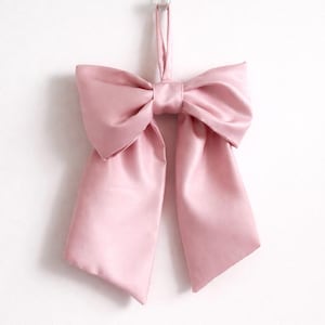 Pode incluir: Um grande laço de cetim rosa claro, pendurado em um gancho adesivo transparente. O laço tem uma forma clássica com um nó central e caudas longas e esvoaçantes. O tecido tem um brilho sutil, em um fundo branco.