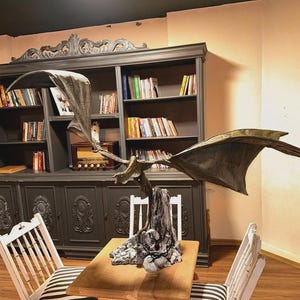 May include: A bronze dragon sculpture with extended wings, mounted on a wooden base. The sculpture is the centrepiece of a small table, with two white chairs featuring black and white striped cushions. A dark grey bookcase filled with books is in the background.