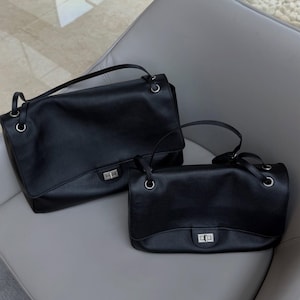 May include: Two black leather handbags of different sizes, each featuring a rectangular flap, a silver-toned metal turn-lock closure, and thin shoulder straps attached to the bag with silver-toned metal grommets and rings.