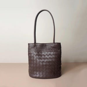 Puede incluir: Un bolso tote marr&oacute;n oscuro con un dise&ntilde;o de cuero tejido. El bolso tiene dos asas largas y una forma rectangular. El bolso est&aacute; hecho de un material de cuero suave y flexible.