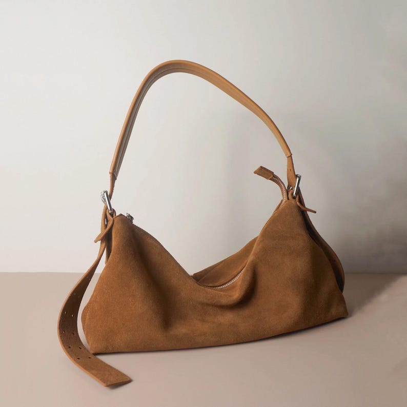 May include: A brown suede shoulder bag with a curved handle and a long adjustable strap. The bag has a zipper closure and a soft, slouchy design. The bag is made of a soft, suede material.
