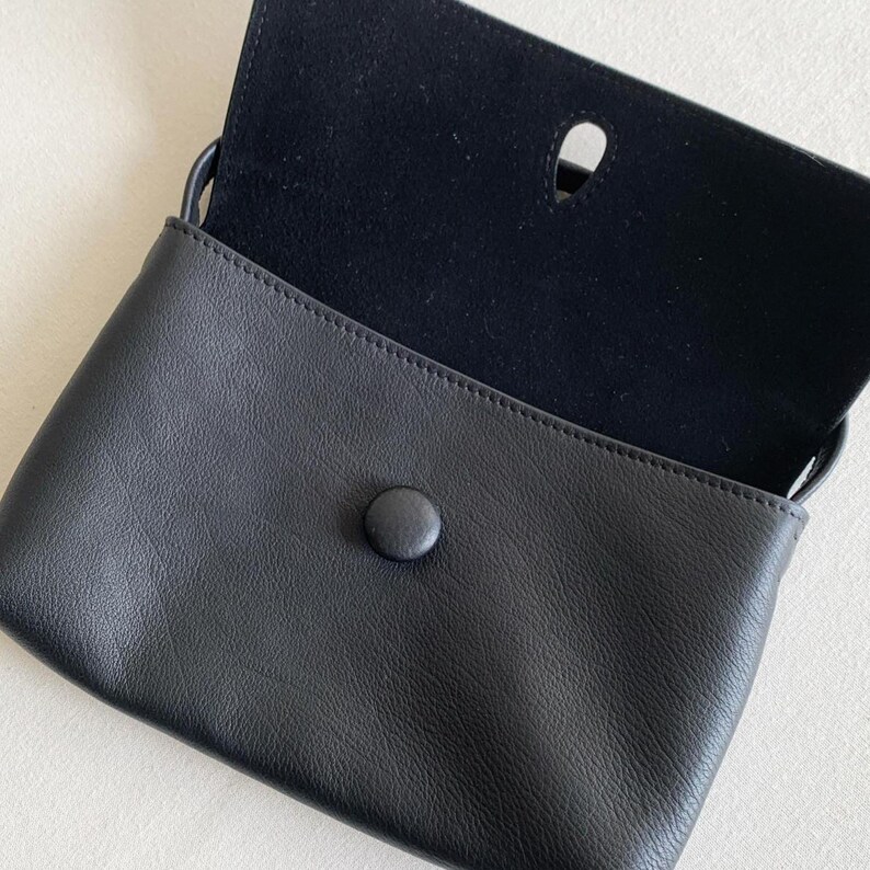 May include: A black leather pouch with a smooth exterior and a textured flap. The flap features a central teardrop-shaped cutout and a single black button closure. The interior of the flap is lined with a soft, dark, matte material.